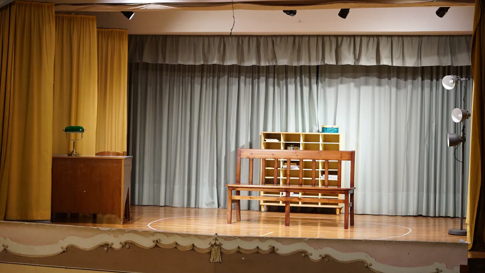 Palco teatrale allestito con panchina, scrivania e luci